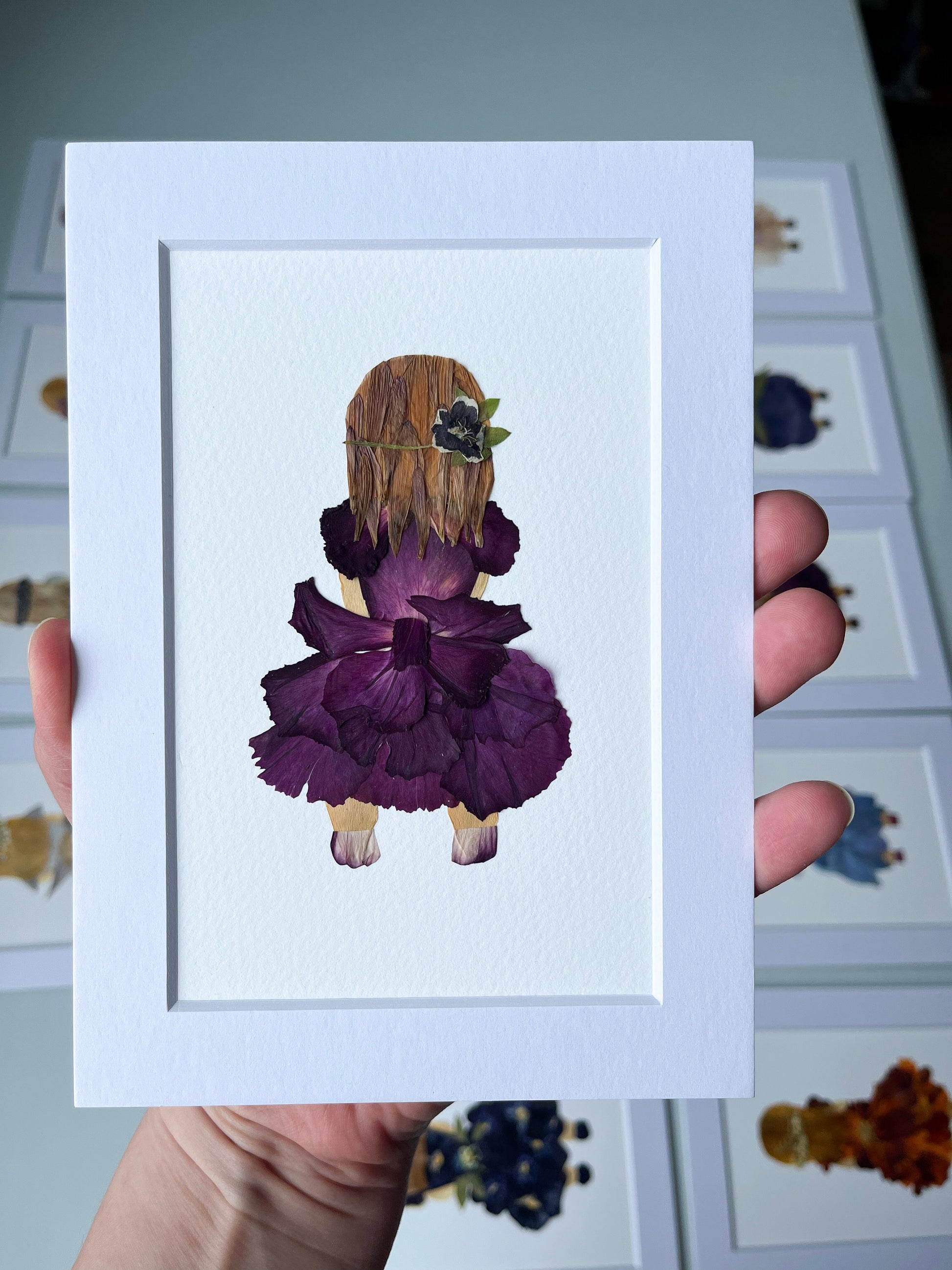Framed artwork of a girl in a purple dress made from pressed flowers, held by a person.